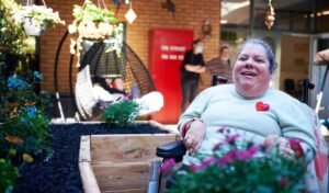 A woman who uses a wheelchair is smiling. Flower beds are in the foreground.