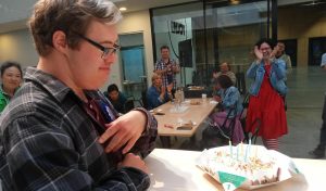 A young man is smiling at a birthday cake while other adults sing.