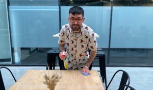 Man with dark hair and glasses is cleaning a wooden table with a spray bottle and cloth.