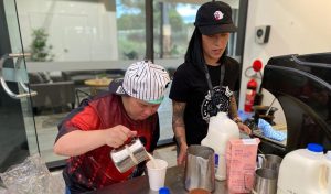 two women, one wearing a white bandana and one wearing a black cap are standing behind a barista bench pouring milk for a coffee